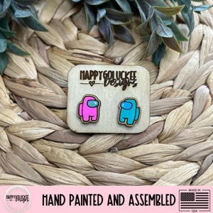 May include: A pair of Among Us character earrings, one pink and one blue, on a wooden backing. The earrings are hand-painted and assembled. The wooden backing has the text "HAPPYGOLUCKEE Designs".