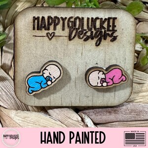 May include: Wooden earrings featuring a sleeping baby design, one in blue and the other in pink, on a wooden display card. The card is engraved with "HAPPYGOLUCKEE Designs" and "HAND PAINTED" is printed below.