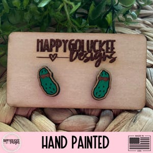 May include: Wooden earrings featuring a pickle design. The earrings are hand-painted and set on a light brown wooden backing. The text "HAPPYGOLUCKEE Designs" is engraved on the wooden backing. The words "HAND PAINTED" are printed below.
