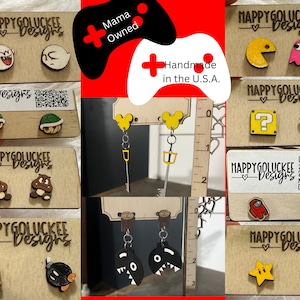May include: A set of handmade wooden earrings featuring various video game characters, including Pac-Man, Ghost, Mario, Luigi, and more. The earrings are displayed on a wooden background with a ruler for scale. The text "HappyGoluckee Designs" and "Mama Owned" is visible on the cards.