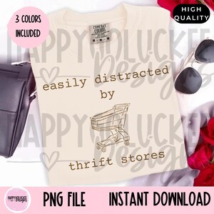 Puede incluir: Camiseta color crema con el texto "easily distracted by thrift stores" y un gráfico de carrito de compras. La camiseta incluye el texto "3 colores incluidos" y "Alta calidad".