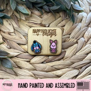 May include: Wooden stud earrings featuring Eeyore and Piglet characters. The earrings are hand-painted with blue, pink, and other colors. The earrings are on a wooden square with the text "HAPPY GOLUCKEE Designs". The bottom of the image says "HAND PAINTED AND ASSEMBLED".