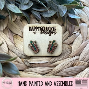 May include: Wooden earrings featuring a brown iced coffee design with a green straw. The earrings are on a wooden rectangular base with the text "Happy Go Lucky Designs". The earrings are hand painted and assembled.
