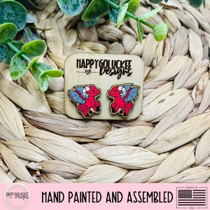 May include: Wooden dragon earrings with red bodies, blue wings, and black outlines. The earrings are hand-painted and assembled. The text "HAPPY GOLUCKEE Designs" is above the earrings. The background is a woven, light brown material.