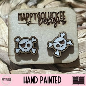 May include: Wooden stud earrings featuring a pirate skull and crossbones design. The earrings are hand-painted with white and brown accents. The earrings are displayed on a wooden card with the text "HAPPYGOLUCKEE Designs". The words "HAND PAINTED" are also visible.
