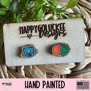 May include: Hand-painted wooden stud earrings. One earring features a blue cat face with yellow eyes, and the other a green plant in an orange pot. The earrings are on a wooden display card with the text "HAPPYGOLUCKEE Designs".