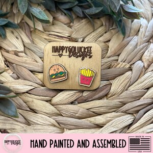 May include: Wooden earrings featuring a hamburger and a carton of fries. The earrings are hand-painted and assembled. The text "HAPPY GOLUCKEE Designs" is above the earrings. The earrings are made in the USA.
