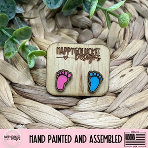 May include: Wooden earrings featuring baby footprints, one pink and one blue, on a light brown square. The earrings are hand-painted and assembled. The text "HAPPYGOLUCKEE Designs" is above the footprints. The background is a woven texture.