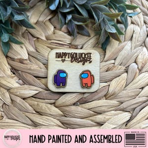 May include: Wooden earrings featuring two Among Us characters, one purple and one orange, with blue visors. The earrings are hand-painted and assembled, as indicated by the text on the image. The earrings are displayed on a woven background.