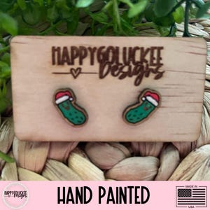 May include: Wooden earrings featuring a green pickle wearing a Santa hat. The earrings are hand-painted and displayed on a wooden card with the text "Happy Go Lucky Designs". The card is on a woven surface.
