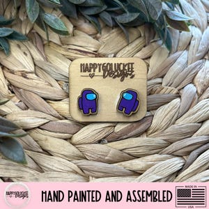 May include: Wooden stud earrings featuring a purple Among Us character with a blue visor. The earrings are hand-painted and assembled. The earrings are displayed on a wooden square with the text "HAPPY GOLUCKEE Designs".