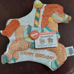 Puede incluir: Un molde para pastel de Wilton en forma de caballo de carrusel con un mensaje de feliz cumpleaños. El molde está decorado con glaseado naranja, amarillo y azul. El molde está diseñado para hacer un pastel de 2 capas.
