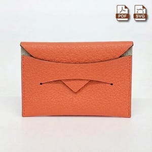 May include: An orange leather card holder with a textured surface. The card holder has a flap closure and a small, triangular accent. The interior is a neutral color. The overall design is minimalist and modern.