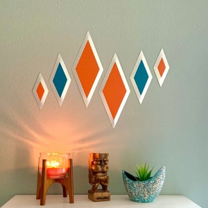 May include: A white cabinet with two drawers, one orange and one teal. The cabinet is decorated with a row of orange and teal diamond shapes on the wall behind it. A wooden tiki statue, a candle holder with a lit candle, and a teal planter with a small plant are on top of the cabinet.