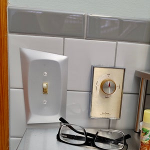 May include: A silver light switch plate with a white toggle switch and two screws. A white knob with a red pointer and the text "off", "heat", "very hot", and "very cool" is mounted on a white tile wall. The knob is labeled "Nu Tone Heat-A-Vent".