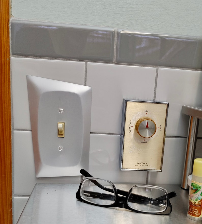 May include: A silver light switch plate with a white toggle switch and two screws. A white knob with a red pointer and the text "off", "heat", "very hot", and "very cool" is mounted on a white tile wall. The knob is labeled "Nu Tone Heat-A-Vent".