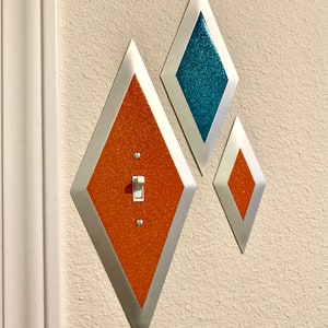 May include: Three diamond-shaped wall plates with a light switch in the center of the largest plate. The plates are silver with orange and blue glitter.