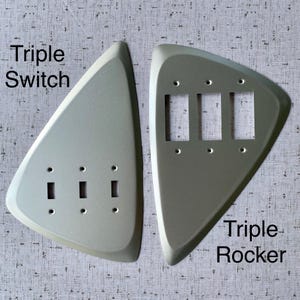 May include: Two silver-coloured, triangular-shaped wall plates. The left plate is labelled Triple Switch and features three small rectangular openings. The right plate is labelled Triple Rocker and features three larger rectangular openings.