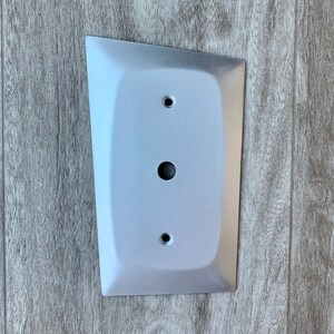May include: A white, rectangular wall plate with a rounded edge and three holes. The plate is mounted on a wood-grain surface.