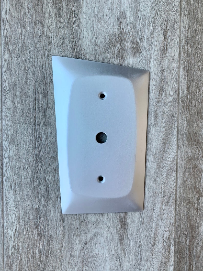 May include: A white, rectangular wall plate with a rounded edge and three holes. The plate is mounted on a wood-grain surface.