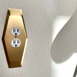 May include: A gold-colored outlet cover with a hexagonal shape and two standard electrical outlets. The cover is mounted on a white wall.