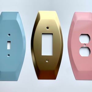 May include: Three decorative wall plates in light blue, gold, and pink. Each plate has a different design: a single light switch, a double light switch, and a single electrical outlet.