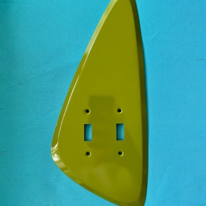 May include: A lime green, teardrop-shaped wall plate cover featuring two rectangular cutouts for toggle switches and four small circular holes for mounting screws. The surface is smooth and glossy, with a distinct asymmetrical, curved silhouette.