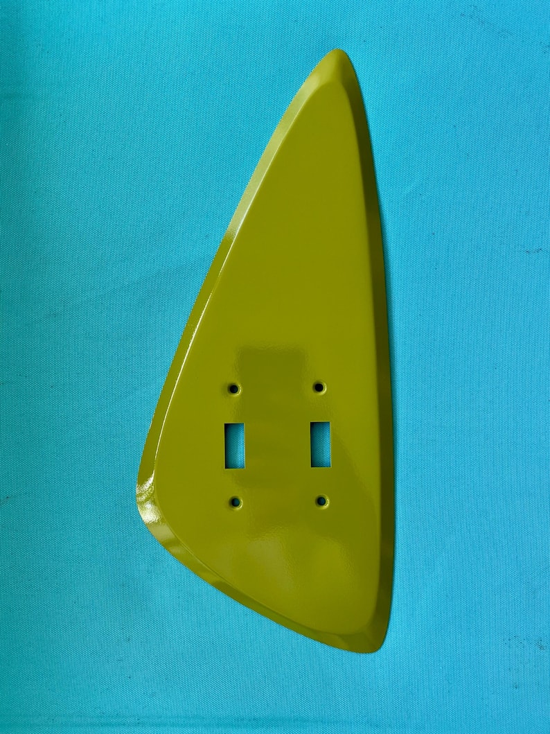 May include: A lime green, teardrop-shaped wall plate cover featuring two rectangular cutouts for toggle switches and four small circular holes for mounting screws. The surface is smooth and glossy, with a distinct asymmetrical, curved silhouette.