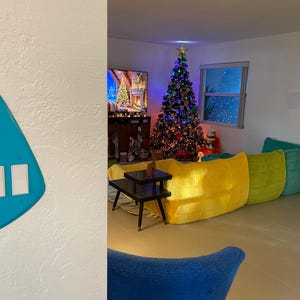 May include: A bright turquoise, asymmetrical, triangular-shaped light switch wall plate featuring two white rectangular rocker switches mounted on a white wall, positioned in the foreground of a room with a Christmas tree and colourful furniture.