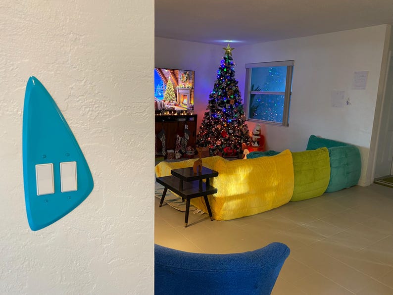 May include: A bright turquoise, asymmetrical, triangular-shaped light switch wall plate featuring two white rectangular rocker switches mounted on a white wall, positioned in the foreground of a room with a Christmas tree and colourful furniture.