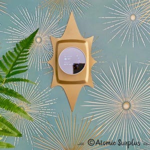 May include: A gold-colored, starburst-shaped wall-mounted thermostat with a round digital display. The thermostat is set against a teal and gold starburst patterned wallpaper. A green fern leaf is visible on the left side of the image. The text "Atomic Surplus" is at the bottom.