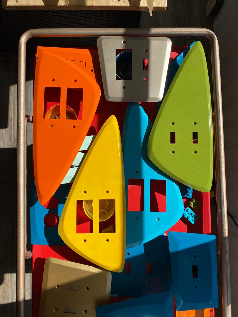 May include: A collection of assorted plastic light switch plates in various shapes and colours, including orange, yellow, lime green, blue, and beige. The plates feature rectangular and square cutouts for switches and mounting screw holes.