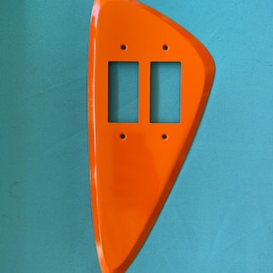 May include: A bright orange, asymmetrical wall plate cover featuring a smooth, glossy finish. This decorative switch plate is designed to accommodate two rectangular rocker-style light switches and includes four small circular screw holes.