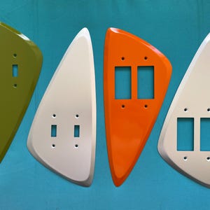 May include: Four asymmetrical, triangular-shaped light switch wall plates arranged in a row. The set includes one olive green plate, one white plate, one bright orange plate, and one white plate, each featuring cutouts for toggle or rocker switches.