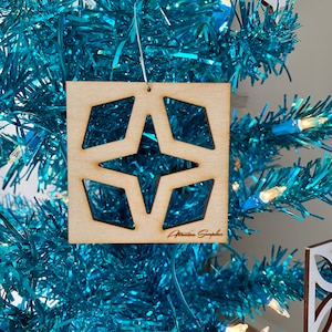 May include: A square wooden Christmas ornament with a star and diamond cut-out design. The ornament hangs on a turquoise tinsel Christmas tree with blue lights. The ornament has the text "Attannie Simple" on it.