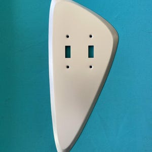 May include: A white, asymmetrical, teardrop-shaped wall plate cover designed for a double toggle light switch. The plate features two rectangular cutouts for switches and four small circular holes for mounting screws, with a smooth, solid finish.