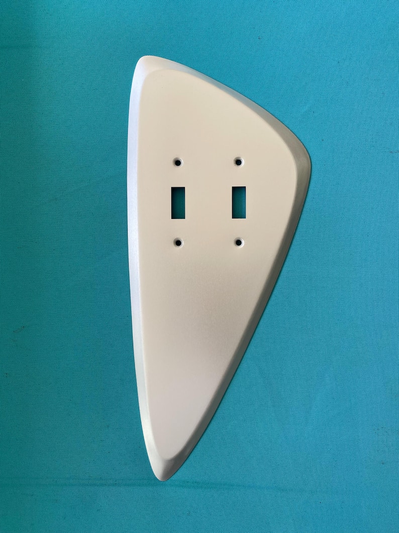 May include: A white, asymmetrical, teardrop-shaped wall plate cover designed for a double toggle light switch. The plate features two rectangular cutouts for switches and four small circular holes for mounting screws, with a smooth, solid finish.