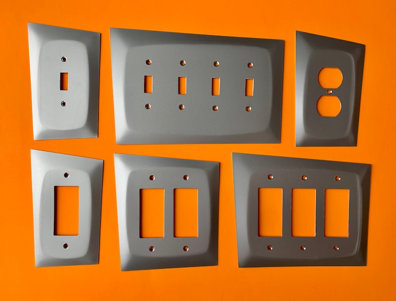May include: Six metal electrical wall plates in a variety of sizes and configurations. The plates are all a light gray color and have a slightly textured surface. The plates are arranged in two rows of three, with the top row containing a single switch plate, a double switch plate, and an outlet plate. The bottom row contains a single switch plate, a double switch plate, and a triple switch plate.