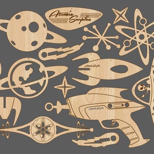 May include: A collection of laser-cut wooden shapes with a retro space theme. Includes planets, rockets, a ray gun, and atomic symbols. The text "Atomic Surplus" and "Astro Probe" are visible on some pieces. The pieces are light brown.