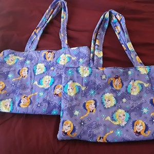 May include: Two purple tote bags with a Frozen theme. The bags feature images of Elsa and Anna from the movie Frozen. The bags have the text "Sisters Forever" printed on them.
