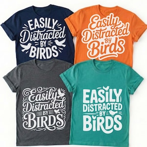 May include: Four t-shirts in navy, orange, charcoal, and teal, each with the phrase "Easily Distracted By Birds" in white script and block lettering, along with bird illustrations. The shirts are short-sleeved.