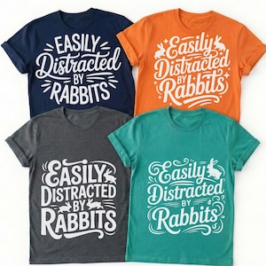 Könnte beinhalten: Vier T-Shirts in Marineblau, Orange, Anthrazitgrau und Türkis. Jedes Shirt hat den weißen Text "Easily Distracted by Rabbits" mit Hasenillustrationen. Die Shirts sind kurzärmlig.