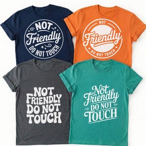 May include: Four short-sleeved t-shirts in navy blue, orange, dark gray, and teal. Each shirt features white graphic text that reads "NOT Friendly DO NOT TOUCH" in various stylized fonts and circular or stacked layouts printed on the front chest.