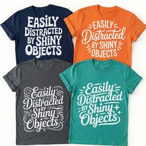 Puede incluir: Cuatro camisetas en azul marino, naranja, gris carbón y verde azulado. Cada camiseta presenta el texto blanco "EASILY DISTRACTED BY SHINY OBJECTS" en una fuente estilizada. Las camisetas tienen mangas enrolladas y cuello redondo.