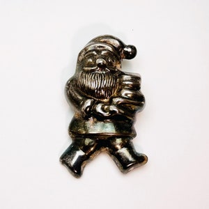 May include: A silver-coloured Santa Claus figurine, shown walking and carrying a sack. The figure has a detailed beard, hat, and boots. The figurine is likely made of metal and is a festive holiday decoration.