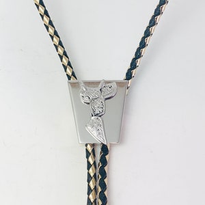 May include: A silver bolo tie with a black and gold braided cord. The tie features a silver charm in the shape of a saddle.