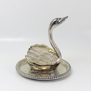 May include: A silver swan ornament with a blue gem eye, on a round, ornate tray. The swan's wings feature feather patterns. A decorative item for display, possibly for a dressing table.
