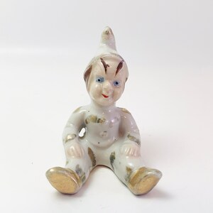 Peut inclure: Une petite figurine en porcelaine blanche d'un lutin assis portant un chapeau pointu. Le lutin a les cheveux bruns, les yeux bleus et un sourire peint. La figurine est décorée d'accents dorés.