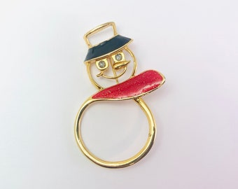 May include: A gold snowman brooch with a dark blue hat and a red scarf. The snowman has two clear crystal eyes and a gold wire frame for the nose and mouth. A festive brooch for the holiday season, perfect for a coat or scarf.