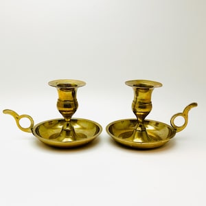 Peut inclure: Deux bougeoirs en métal doré avec un petit plat et une poignée. Les bougeoirs sont sur un fond blanc.
